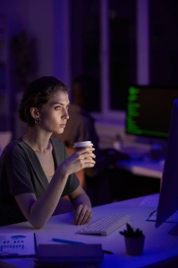 Modern genç kadın gecenin geç saatlerinde karanlık bir ofiste oturuyor kahve içiyor ve bilgisayar ekranına bakıyor.