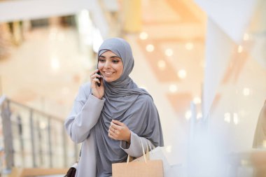 Orta Doğulu zarif bir kadının telefonla konuşan ve alışveriş merkezinde dikilirken başörtüsü takan portresi.
