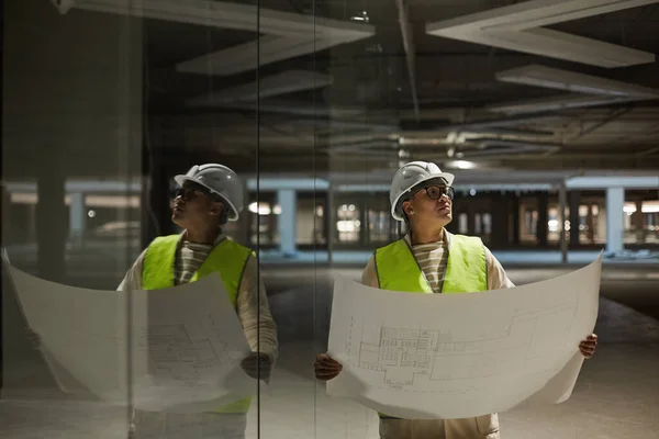 Waist up portrait of female engineer holding blueprints while inspecting base of building at construction site, copy space