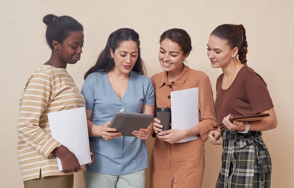 Multi-ethnic group of four young businesswomen looking at digital tablet while standing by wall in office