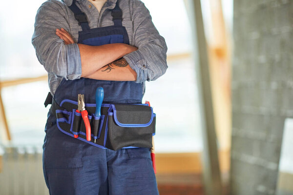 Cropped portrait of contemporary female worker standing with arms crossed with focus on tool belt, copy space