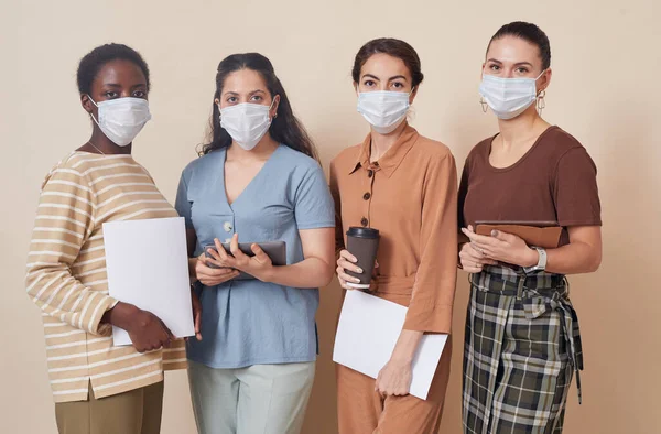 Multi-ethnic group of four young businesswomen wearing masks and looking at camera while standing by wall in office