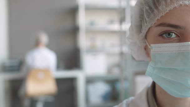 近半数的年轻白种人女性坐在实验室时 头戴一次性医疗帽 戴着口罩 面带微笑地看着相机 — 图库视频影像