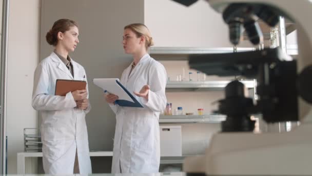 生化学の研究室で一緒に立っている医療を身に着けている若い女性科学者のペアの焦点のショットをラック通信 — ストック動画