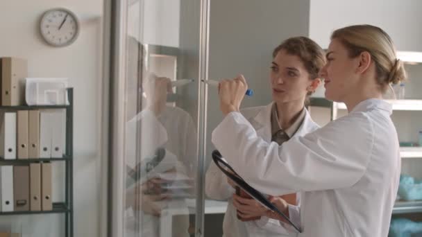 两名身穿医疗服的白人妇女在实验室站在一起 在玻璃墙上画化学配方的中片照片 — 图库视频影像