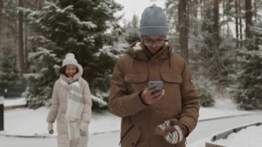 Orta boy Afrikalı bir adamın elinde cep telefonuyla buz pateni pistinde dururken kıvırcık saçlı bir kadının ona yaklaşıp telefon ekranına baktığı bir fotoğraf.
