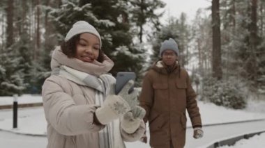 Açık havada buz pateni pistinde elinde akıllı telefonuyla duran ve arkasında Afro-Amerikan erkek arkadaşı buzla selfie çeken orta boy melez bir kadın resmi.