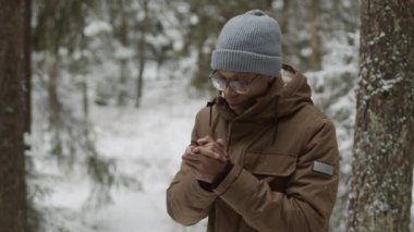Siyah ceketli, şapkalı ve gözlüklü genç bir adamın kış ormanında durup sıcak nefesiyle dondurucu ellerini ısıttığı orta boy bir fotoğraf.