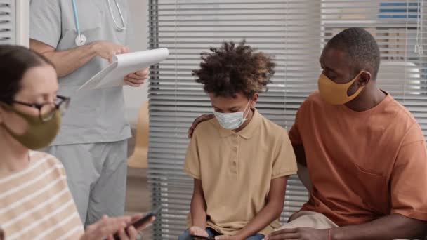 中長期の多様な人々のマスクを着て 待合室に座っている 兄はアフリカの学齢者の少年を安心させました 彼のオフィスで彼を招待認識できない医師 — ストック動画