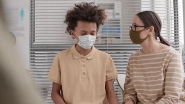 Bel yukarı, kahverengi saçlı beyaz kadın ve Afrika 'da okul çağında yüz maskesi takan, ofiste oturan çocuk. Anne oğluyla konuşuyor.