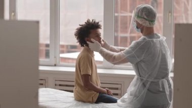 Tanınmayan, virüs korumalı bornoz, eldiven ve yüz maskesi takan orta boy bir doktor, tıbbi kanepede oturuyor, Afro-Amerikan okul çağındaki çocuğu muayene ediyor, konuşuyor, omzunu okşuyor.