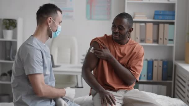若いアフリカ人男性の中長期医療室に座って 肩の上に綿のパッドを押すと 話して 予防接種やバンピング拳のための白人男性医師に感謝 — ストック動画