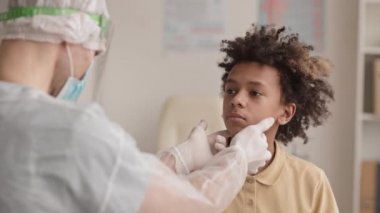 Afro-Amerikan okul çağındaki çocuğun omuzları üzerinde virüs koruması ve eldiven giyen tanınmayan bir doktor tarafından salgı bezleri kontrol ediliyor.