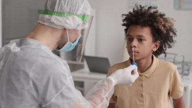 Okul çağındaki Afrikalı bir çocuğun omuzlarının üstünde tanınmayan bir tıp işçisinin analiz için pamuklu çubukla burnundan örnek almasına izin veriyor. Hasta gülümsüyor ve başını sallıyor.