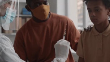 Orta ölçekli, Afrika 'da okul çağında, maske takan ve vurulmadan önce onu destekleyen bir çocuk. Doktorun yüz kalkanı takıp şırıngaya bastığını, sıvının fışkırdığını...