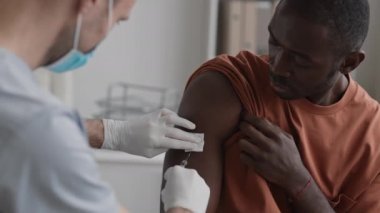 Genç Afrikalı bir adamın göğsü tıbbî ofiste otururken gömleğinin kolunu kaldırmış, tanınmayan bir doktorun hastanın omzuna pamuk pedi bastırıp aşı yaparken.