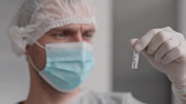 Tıbbi maske takan, tek kullanımlık şapka takan ve eldiven takan, COVID-19 aşısıyla ampul tutan bulanık laboratuvar çalışanlarının yakın çekimi.