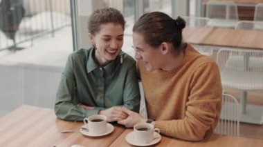 Orta boy kıvırcık saçlı beyaz erkek ve kadının şık kıyafetler giyip restoranda buluştuğu bir fotoğraf. Pencereden bakıp gülüyorlar.
