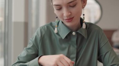 Kafe masasında oturan, çay kaşığıyla kahve karıştıran ve kameraya bakıp gülümseyen kıvırcık saçlı, güzel beyaz bir kadın.