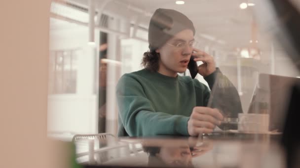 中等比例的年轻嬉皮士男子穿着便服和眼镜坐在笔记本电脑前的咖啡店里 在讲手机 — 图库视频影像
