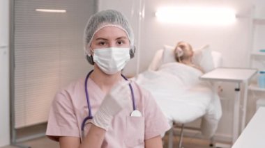 Hasta koğuşundaki beyaz kadın doktorun göğüs röntgeni, önlük, tek kullanımlık şapka, boynuna steteskop, maske takmak, kameraya bakmak. Uzun bir günün ardından sağlık çalışanının portresi