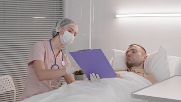 年轻女医生戴着刷子 一次性帽子 手套和面罩 坐在生病的白人男子床边 给他看医生签名 然后走人 — 图库视频影像
