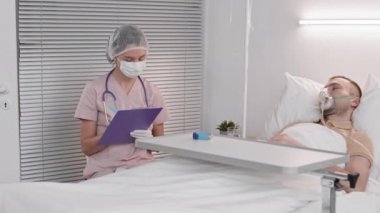 Hastane yatağında yatan oksijen maskeli bir adamın koğuşunda duran beyaz kadın doktor. Sıcaklığını ölçerek, hasta kaydına sağlık belirtileri kaydeden bir doktor.