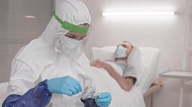 Tam kaplamalı tehlikeli madde giysisi ve yüz kalkanı takmış, ampulden şırıngayı doldurmuş, hastane odasında dikilmiş, sonra da doktor dönüp hastaya bakıyor.