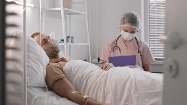 病院のベッドに寝そべっている酸素マスクの男の方に立っている薄ピンクのスクラブを身に着けている若い白人看護師のチェストアップ 患者の記録にメモを取り体温を測定する医療従事者 — ストック動画