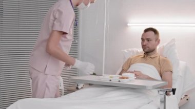 Tek kullanımlık şapka, maske, eldiven takan, beyaz bir adama yemek getirip hastane yatağında yatan, sonra da hastaneden ayrılan bir hastaya teşekkür ederken orta boy kadın hemşire fotoğrafı.