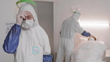 Beyaz tehlikeli madde giysisi giyen, uzun bir günden sonra yüz koruyucusunu ve maskesini çıkaran, kameraya bakan, arka planda hasta olan, bulanık bir meslektaşı olan beyaz kadın doktor.