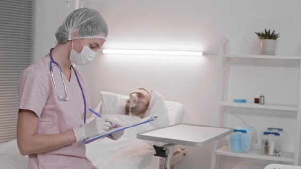 中等年龄的女护士 脖子上戴着刷子 一次性帽子 手套和听诊器 头戴氧气面罩 坐在病人病房里睡觉 填写数据 然后看着摄像机 — 图库视频影像