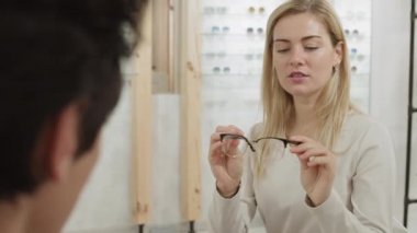 Optisyen dükkanında önünde oturan genç, sarı saçlı kadına farklı gözlük gösteren tanınmayan bir optometristin dikiz görüntüsü.
