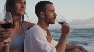 Mavi gözlü kızın arka taraftan yakışıklı erkek arkadaşına sarılışının yan görüntüsü. Rüzgarlı okyanus kıyısında kırmızı şarap içen çift.
