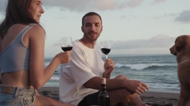 美しい白人の少女と彼女の若いボーイフレンドのアークショット 座って笑って 赤ワインとグラスをクリックし 目を見て 飲んでいます 海岸でロマンチックなピクニックをしているカップル — ストック動画