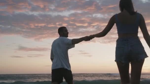 若いロマンチックなカップル保持手の低角度リアビュー 巨大なウエスト 美しい夕日の海沿いの海岸に沿って歩く — ストック動画