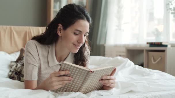 暗い髪の若い白人女性のベッドの上で胃の上に横たわるのSteadicam 本を読んで その後を見上げ それについて考え 笑顔で 物語を楽しむ — ストック動画