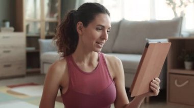 Çekici, beyaz, spor bluz giyen, bulanık oturma odasında oturan, tablet bilgisayardan arayan, el sallayan, gülümseyen, konuşan, su şişesini gösteren genç bir kadının göğsü.