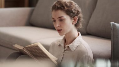 Orta ölçekli, güzel, beyaz bir kadın kanepede oturuyor, kitapta komik hikayeler okuyor, gülüyor ve kameraya bakıp gülümsüyor.