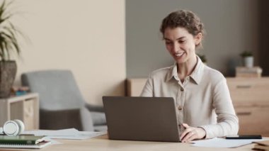 Orta ölçekli, neşeli, beyaz bir kadın ofis masasında oturuyor, bilgisayarını video görüşmesi için kullanıyor, gülümsüyor, konuşuyor, selamlıyor, el sallıyor.