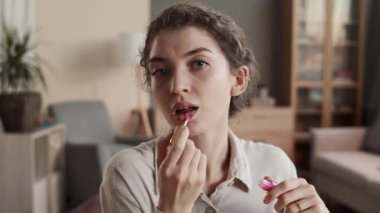 Odasında oturan, dudak parlatıcısı süren, kameraya bakıp gülümseyen beyaz bir kadının göğsünü kabartan pozu.