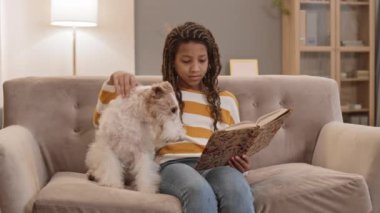 Orta yaşlı Afrikalı kız kitap okuyor kanepede oturuyor, konuşuyor ve köpeğini okşuyor.