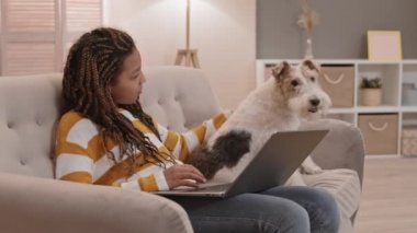 Oturma odasında kanepede oturan, dizüstü bilgisayar kullanan ve Tel Tilki Teriyer köpeğini okşayan okul çağındaki Afrikalı kız çocuğunun orta boy yan görüntüsü.