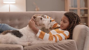 Dokuz yaşındaki Afro-Amerikan bir kızın kitabı bir kenara bırakıp evcil köpeğini okşaması orta uzunlukta bir bakış açısı. Sahibi ve Wire Fox Terrier kanepede uzanıyor.