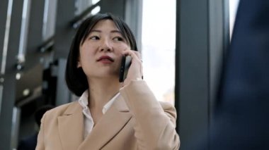 Beyaz ceketli ve gömlekli Asyalı genç bir iş kadınının ofis salonunda dikilip telefon görüşmesi yapması.
