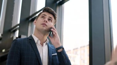 Kafkasyalı genç iş adamının düşük açılı görüntüsü. Kareli mavi takım elbise giymiş. Ofis salonunda duruyor ve telefonla konuşuyor.