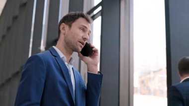 Orta yaşlı, beyaz iş adamı mavi takım elbiseli bir adamın ofis salonunda dikilip telefon görüşmesi yaptığı orta boy bir fotoğraf.