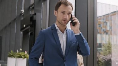 Orta yaşlı Kafkasyalı iş adamının mavi takım elbise giyip ofis koridorunda telefon konuşması yaptığı orta boy bir fotoğraf.