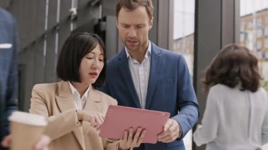 Medium shot of Asian businesswoman and her male Caucasian coworker wearing formalwear going together along office hall, talking and using digital tablet woman holding in hands