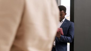 Panning of good-looking African businessman standing in office hall with tablet in hands then his male Caucasian colleague approaching, shaking his hand and starting a chat
